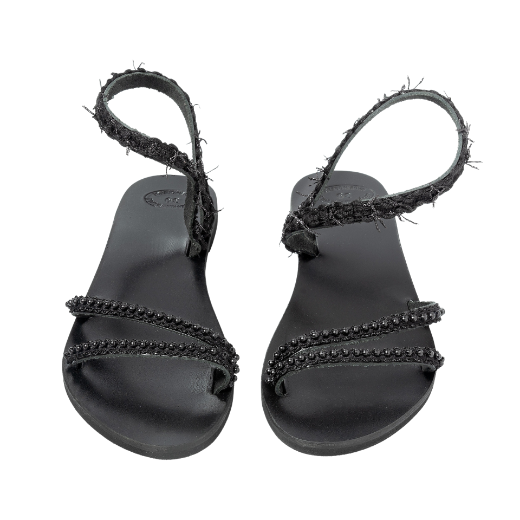 A black sandal with a single textured strap wrapping around the ankle and two thin upper straps, all featuring small decorative elements.
